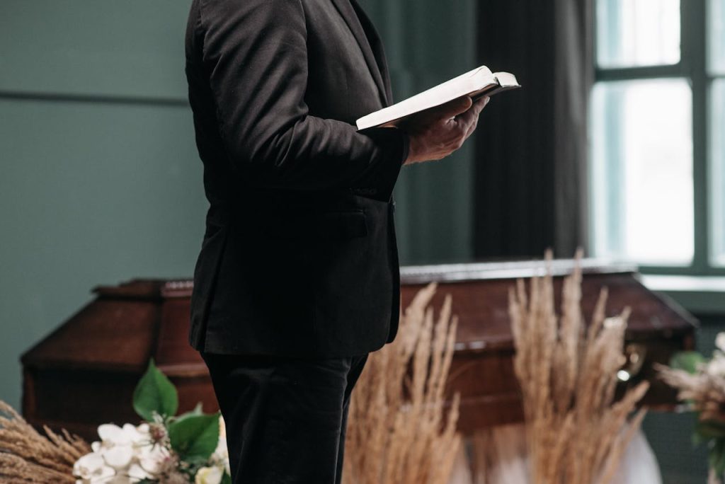 A pastor in a black suit holding a book during a funeral ceremony indoors.